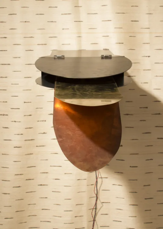 Mounted on fabric, strips of brass and copper extend from the semicircle of a segmented metal box. Wires hang beneath the object.