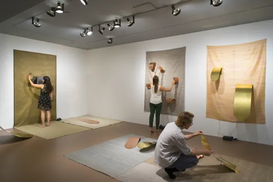 Visitors manipulate gold and copper colored metal objects placed on fabric on the floor and walls. Several objects are tongue-like.