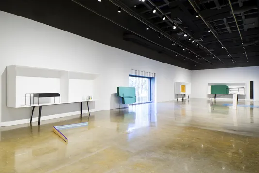 A number of artworks are on display in white shelving units, while other artworks are on the floor and across a picture window.