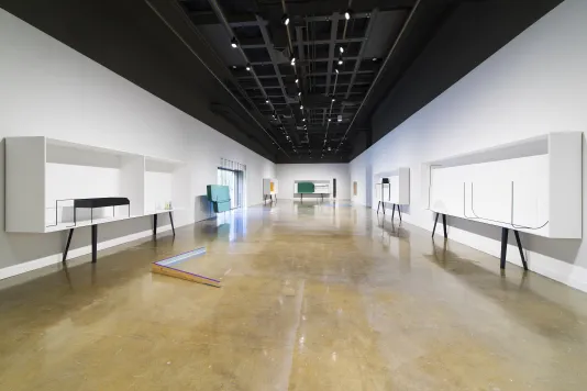 Long view of gallery, showing white display cases with metal legs on right, left and rear walls. V-shaped sculpture on floor.