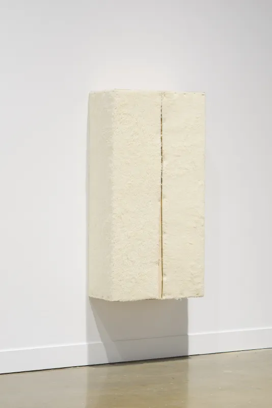A wall-mounted, cream colored sculpture with an orange line down the middle casts a shadow on the wall and floor.