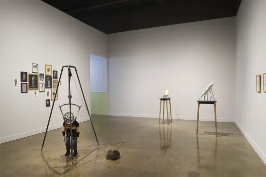 A mix of collages hang, arranged closely together, a metal tripod traps a small figure, and 2 small tables hold objects.