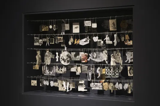 Five rows of paper cutouts hang on strings within a large display case, against a black background.