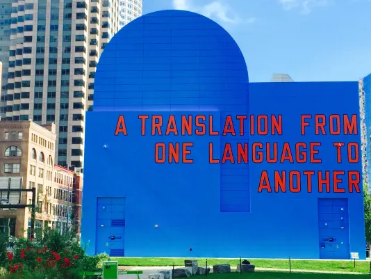 “A Translation From One Language To Another” painted in red and outlined in black on a blue building façade in the cityscape