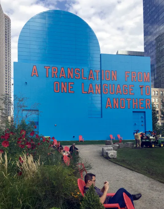 Man in red chair in the park in front of the blue building façade with “A Translation From One Language To Another” in red.