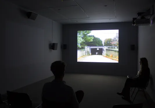 Visitors sit in a darkened gallery looking at a screen showing the entrance to an underpass. A darkened screen is to the left.