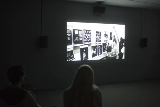 Visitors sit in a darkened gallery looking at a mounted video screen showing people looking at black and white photographs.