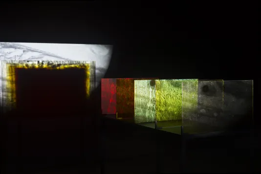 Black and white, green, red images projected onto and through 4 glass panels of the table sculpture fall onto a dark wall.