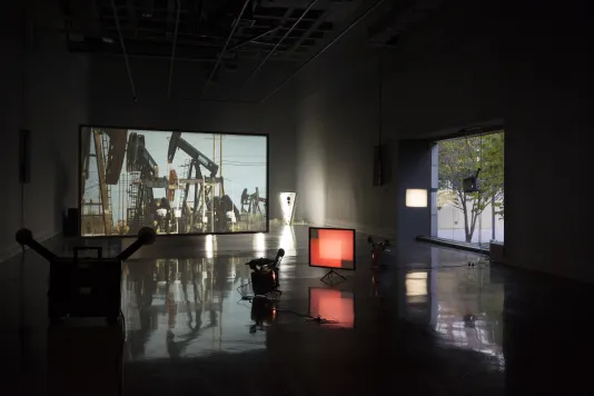 Projectors cast red rectangles on a small foreground screen, a large screen projection of oil drilling rigs in back.
