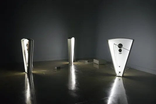 3 triangular sculptures, each internally-lit and fitted with 35mm film and spools, stand before a corner of the dark gallery.