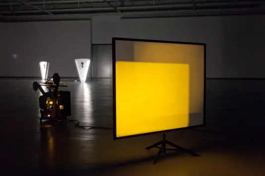 Seated on the floor of a dark gallery, a projector casts overlapping yellow rectangles on a translucent screen.