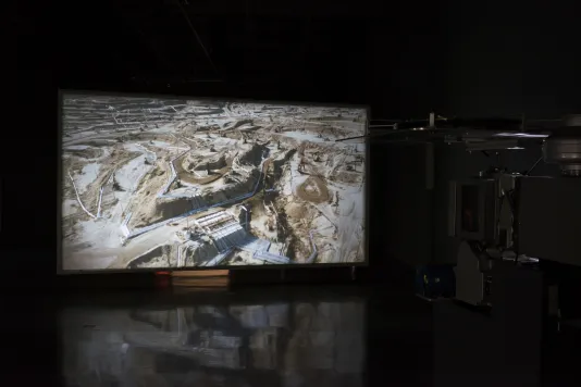 Aerial view of a brown and white excavated landscape is projected on a large screen, next to the large black projector.