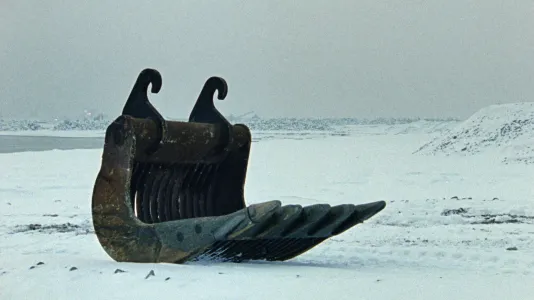 A large rusted, metal excavator rake sits isolated in a white-gray, snow-covered, coastal landscape.