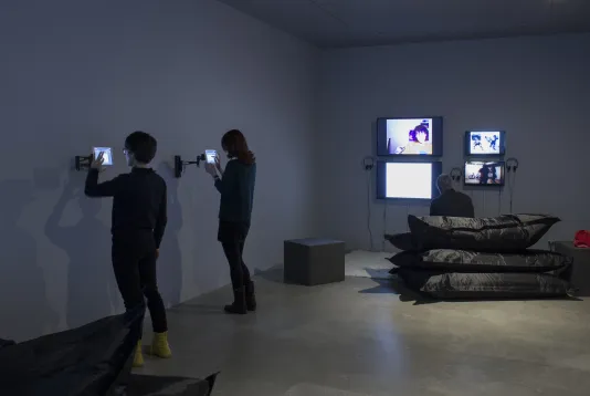 2 visitors interact with wall-mounted iPads while a visitor sits on beanbags watching 4 wall-mounted monitors.
