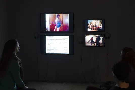 2 visitors watching videos on 4 wall-mounted monitors in a dark gallery.