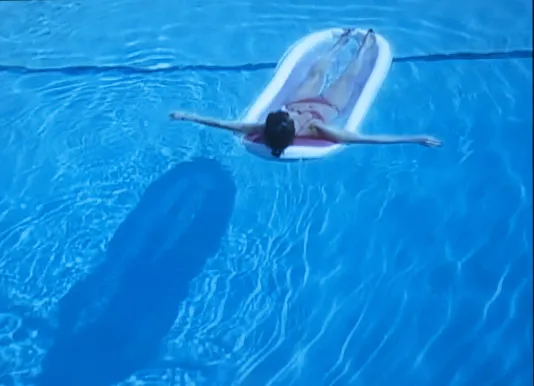 A young woman, clad in pink bikini, with her arms stretched out, float on the bright blue water of a swimming pool.