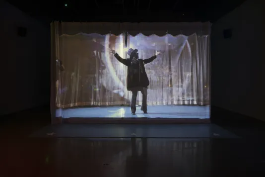 In a darkened space, a projected video of a man in lights, onstage, wearing a top hat and performing with arms outstretched