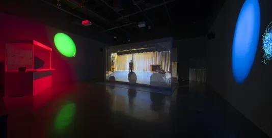 A darkened space with a video screen in the center, large blue and green oval spotlights, and a small bar bathed in red light 