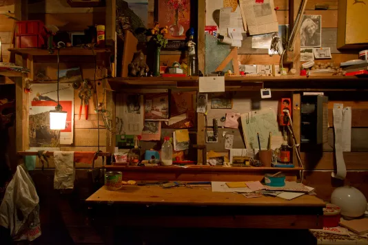 A wood slatted wall, wood table and shelves filled with tools, papers, pencils, pictures, a hanging lamp, on with a warm glow