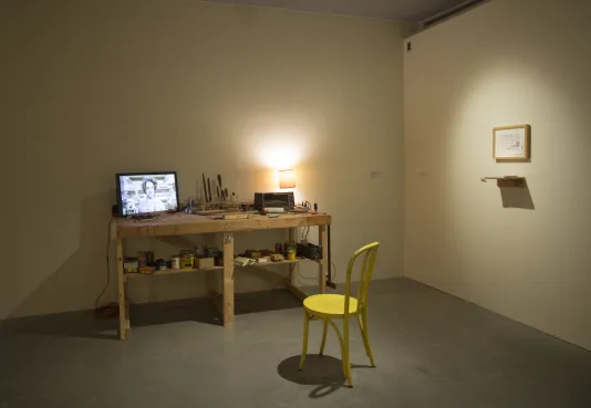Installation view of a work bench with a monitor, tools, a lamp, a yellow chair, a framed piece and shelf on the wall
