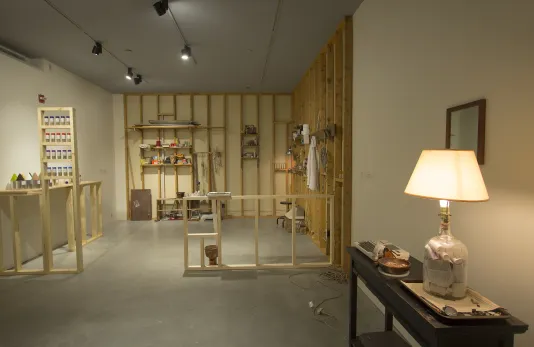 Installation view of a space with wood framing, shelves, a spice rack, found objects, and a narrow table with a lighted lamp