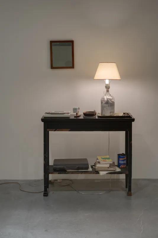 A small table with a lighted lamp, a bowl, computer, a briefcase, coffee can and books, a framed mirror on the wall above