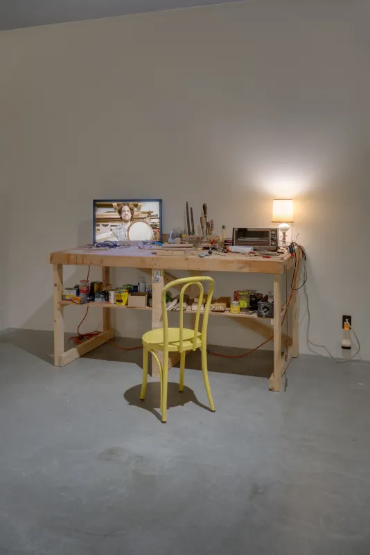 A work bench with a monitor, tools, a toaster oven, a lamp with a warm glow, and a yellow wood chair in front