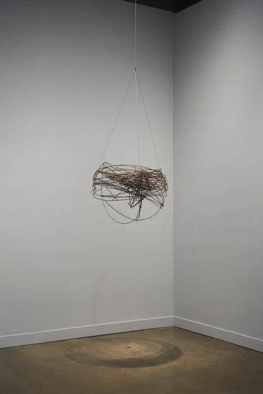 A spherical, brown-bronze-color metal-wire sculpture hangs from the ceiling before white walls in a corner of the gallery.