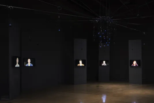 5 monitors each mounted on a black column simultaneously broadcast an individual speaker pitching a scam in a dark gallery