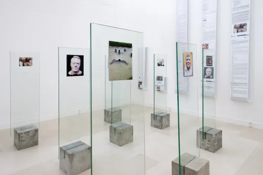 Color photographs mounted on glass panels on concrete bases in the foreground, photos and text on rolls hang on side wall.
