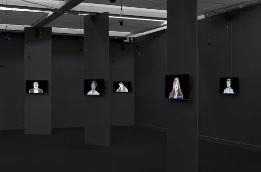 5 monitors each mounted on a black wall show individuals speaking in a darkened gallery.