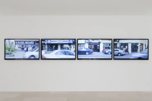 4 monitors, mounted on a white wall, show silver cars driving by in a streetscape with store fronts and pedestrians.
