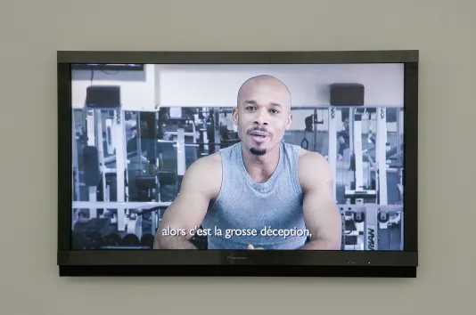 Wall-mounted monitor show a muscular young man in gray tank-top in a gym with the caption “alors c’est la grosse déception,”.