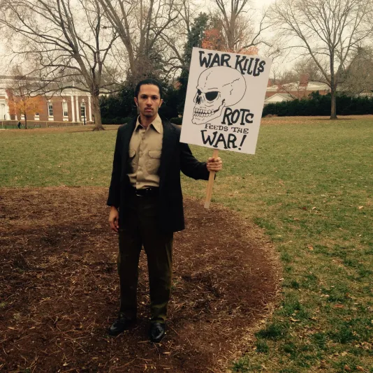 A man in a suit stands with a protest sign that reads &quot;War Kills. ROTC feeds the war!&quot;