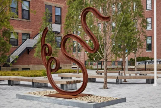 Matt Johnson's Untitled(Swan) sculpture of a warped bent metal train track is pictured in front of MIT campus buildings.