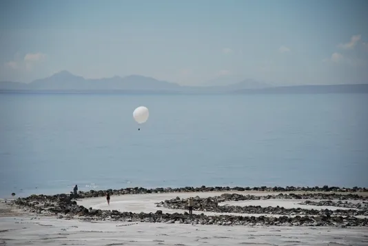 A white balloon floats over a spiral jetty, beach, and ocean.