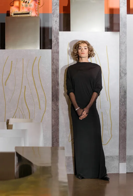 Actor with blond curly hair and wearing black floor-length dress stands before gray façade inscribed with gold drawings.