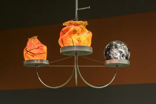 Metal chandelier with 2 orange flame-shaped and 1 black-and-white lit glass orbs hangs in front of rust-color gallery wall.