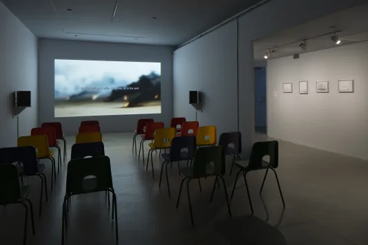 A darkened gallery with projected video at the front, chairs, speakers, and four framed drawings hanging in adjacent space