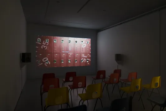 A darkened gallery space set up like a theater with speakers, chairs and a projected video showing two rows of red lockers