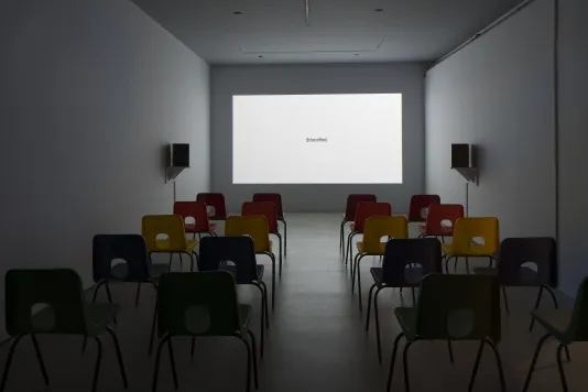 A space with chairs, speakers, a large wall projection of all white, with the word 'intensifies' centered and in parentheses