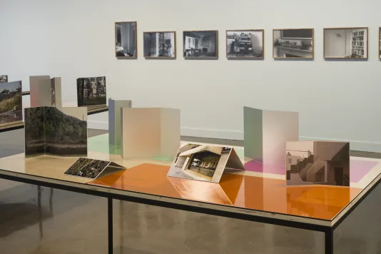 Installation view of two table tops with photographs and color gels, and a row of framed b+w photographs on the wall in back