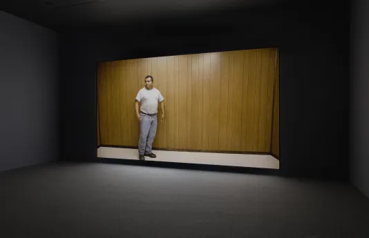 Installation view of video with a man in a t shirt in front of a wood paneled wall, looking out of the frame to the left