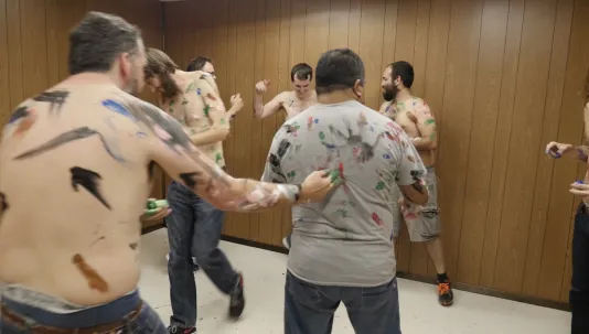 A group of men most without shirts, with painted marks on their torsos. One man reaching out to paint on another’s t-shirt