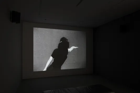 Black and white video projected in a dark room displays woman in black from behind, stretching her right arm in a dance pose.