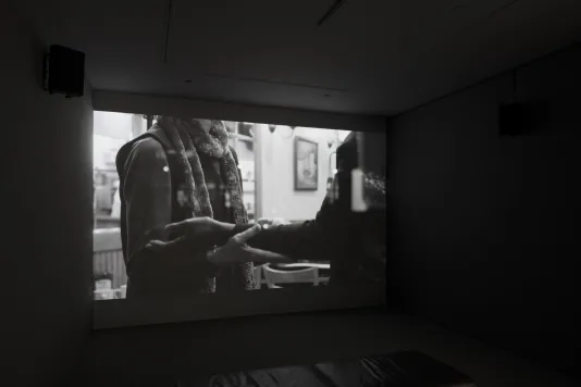 Dark room with video projection of a person's torso, arms bent at right angle, palms up, supporting the arms of another.