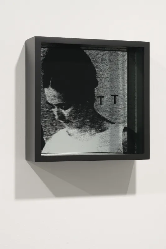 Framed artwork of a woman facing left, head bowed, a letter T on the top glass casts a shadow onto the grainy image.