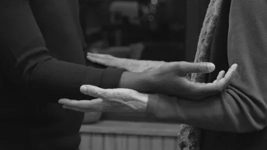 Video still of the arms and hands of a young and old person, forearms out and palms up, resting on top of each other’s arms.