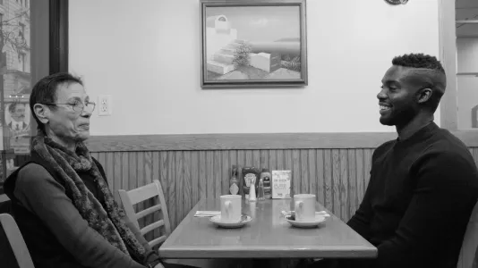 Black and white video still of a seated old woman and a smiling man, facing one another at a cafe table set with coffee.