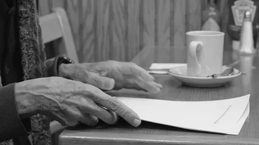 Still closeup of a pair of hands hovering above a cafe table. The fingers of the hand in front bend around sheets of paper.